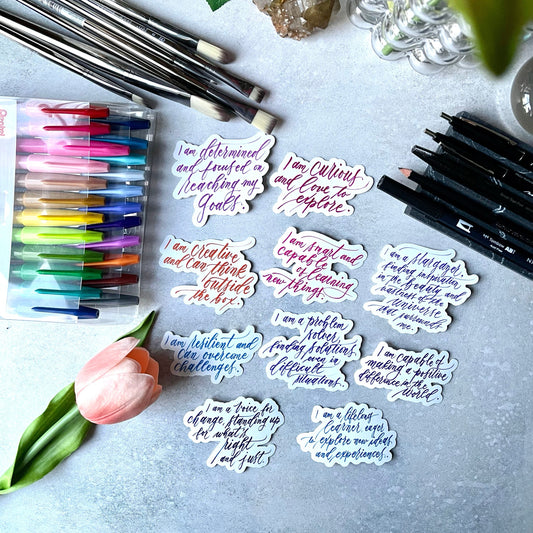 Sticker set of vinyl decal stickers with motivational quotes and inspirational affirmations in colorful calligraphy on a flat lay view of a desk with markers, paint brushes, and tulips on it.