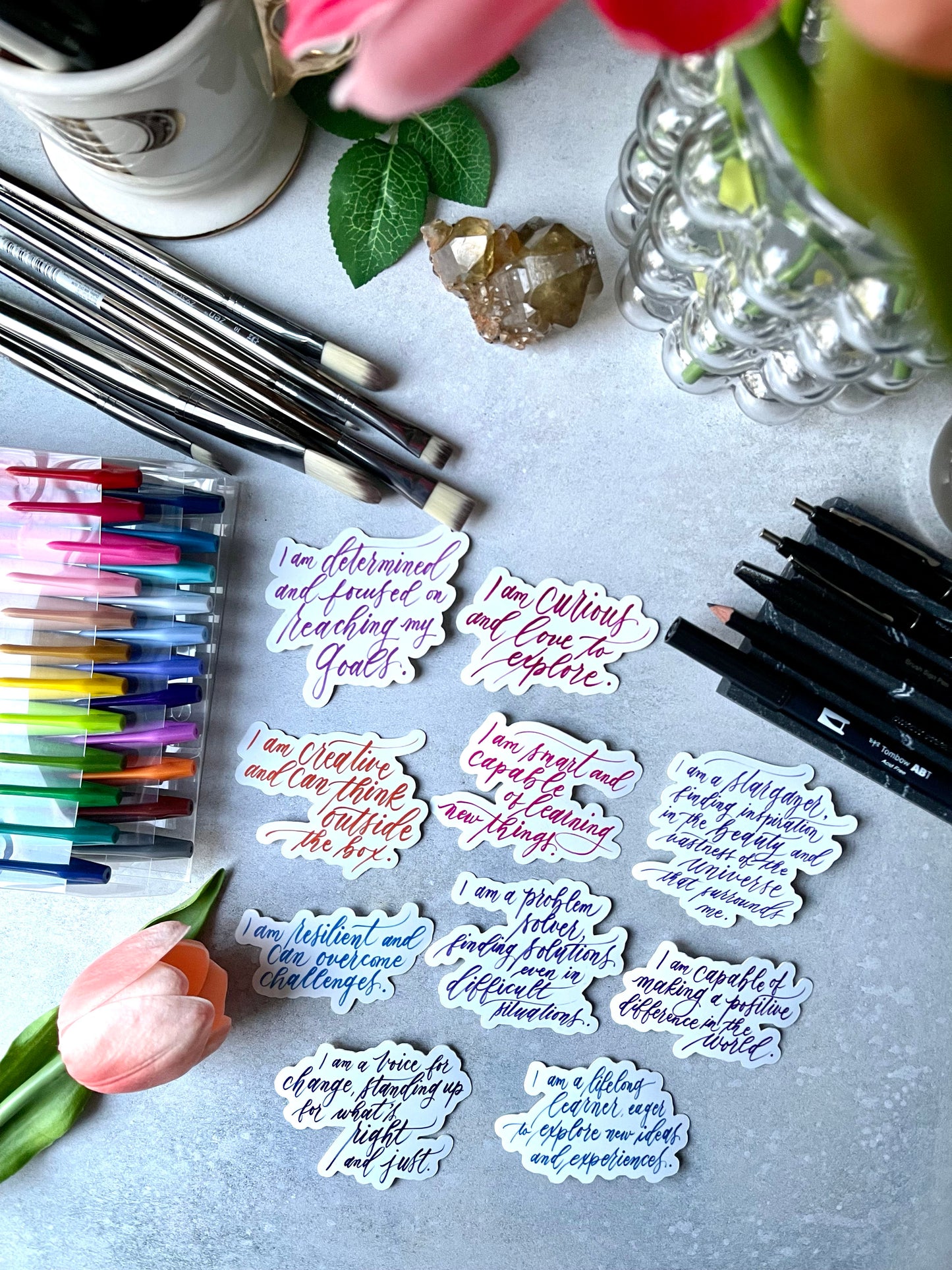 Set of vinyl decal stickers with motivational quotes and inspirational affirmations on a flat lay desk top with colorful markers, a tulip, and crystals.