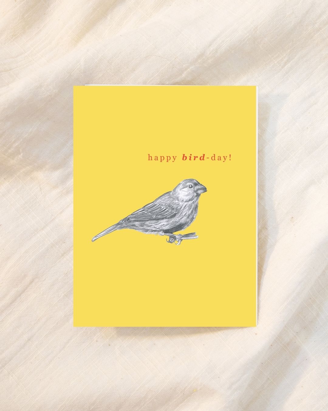 A yellow greeting card with the text 'happy bird-day!' and a sketch-style bird illustration, photographed on a light beige fabric background, hand-sketched for bird lovers and bird watchers.
