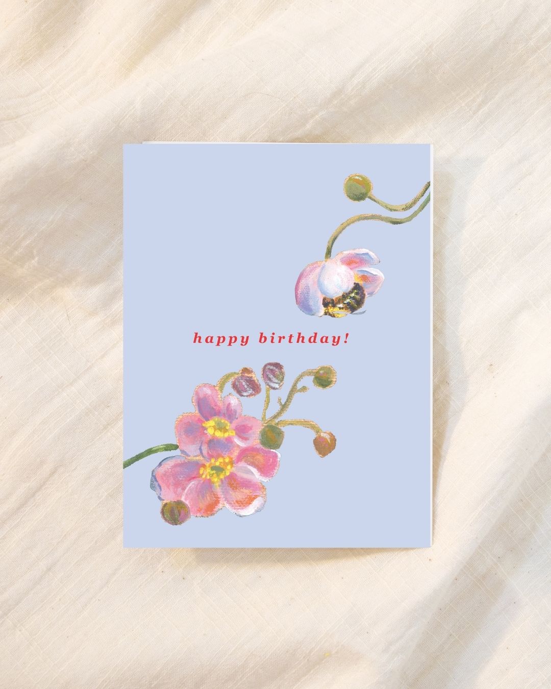 A square birthday card with a light blue background, featuring pink flowers and green stems with a small bee illustration, and the words 'happy birthday!' in red. The card is photographed flat on a cream-colored fabric background.