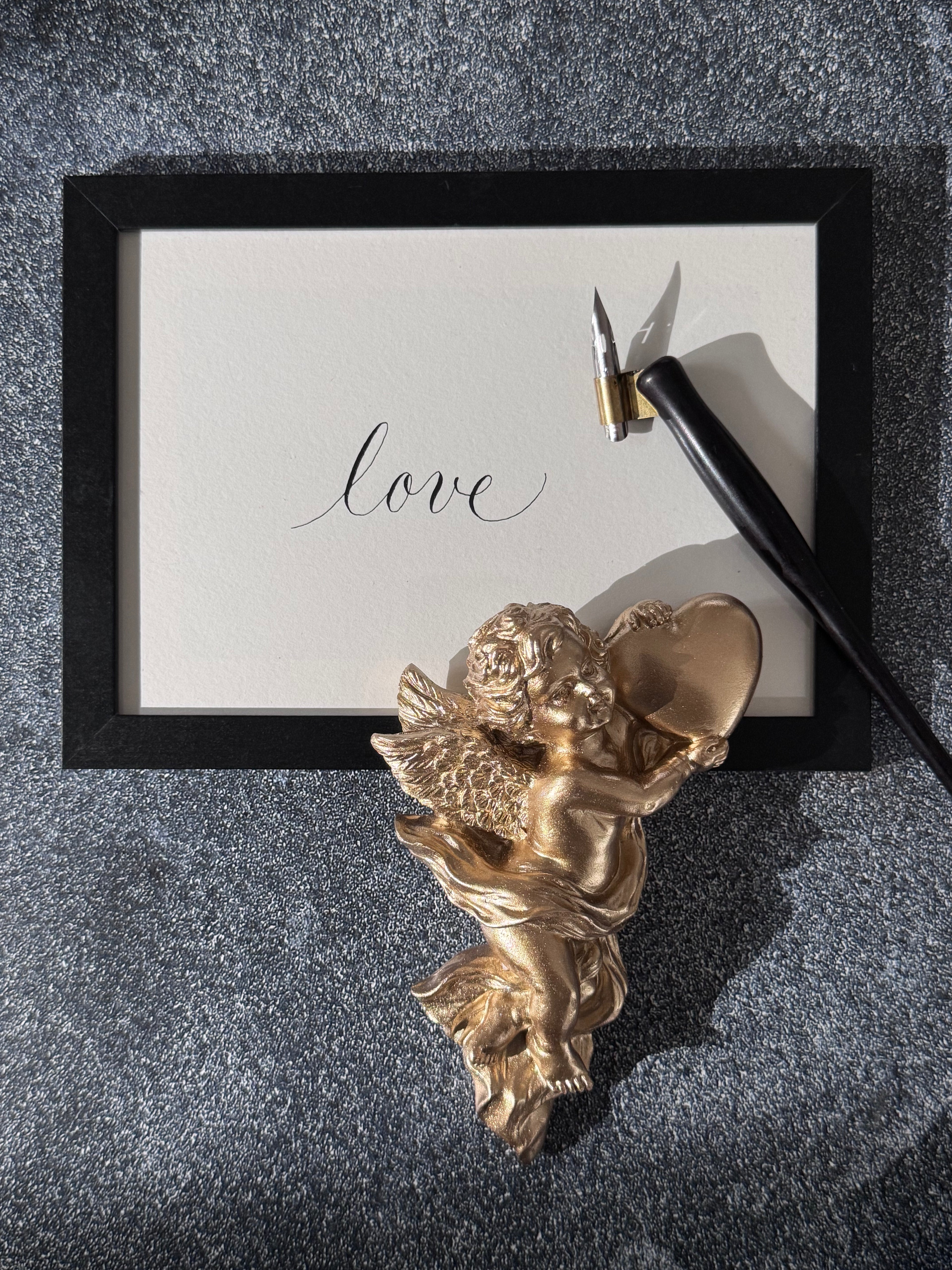 Gold cherub figurine with a pointed ink & nib pen on a textured surface with the word  "love" written in calligraphy