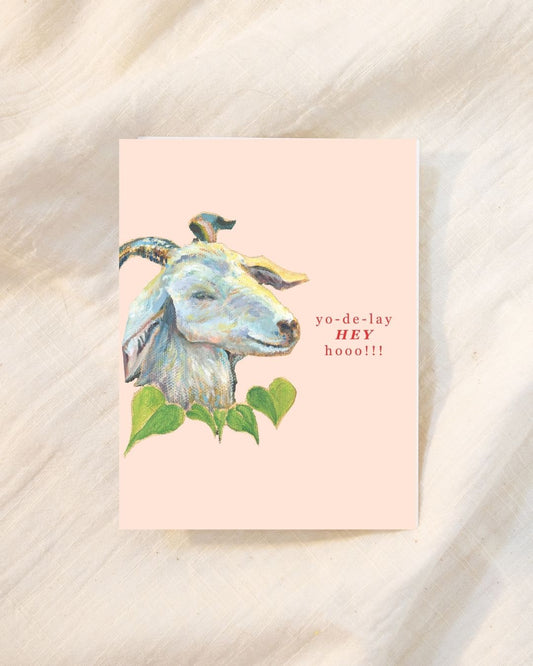 A rectangular greeting card with a pale pink background, featuring a painted goat head with green leaves and the text 'yo-de-lay hey hooo!!!' in red, placed on a soft cream fabric surface.